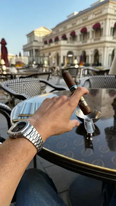 A hand holding a Dominium cigar at an outdoor café table, an ornate European-style building behind