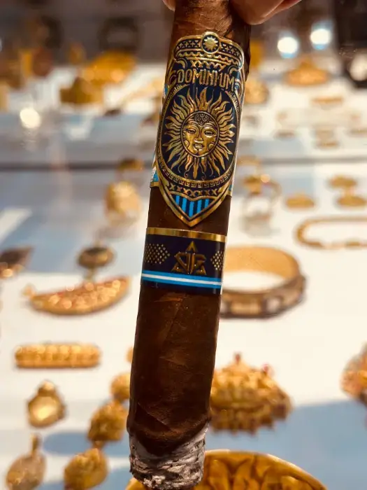 Close-up of a Dominium Primera cigar held against a table scattered with gold jewelry, at a gold souk in Dubai