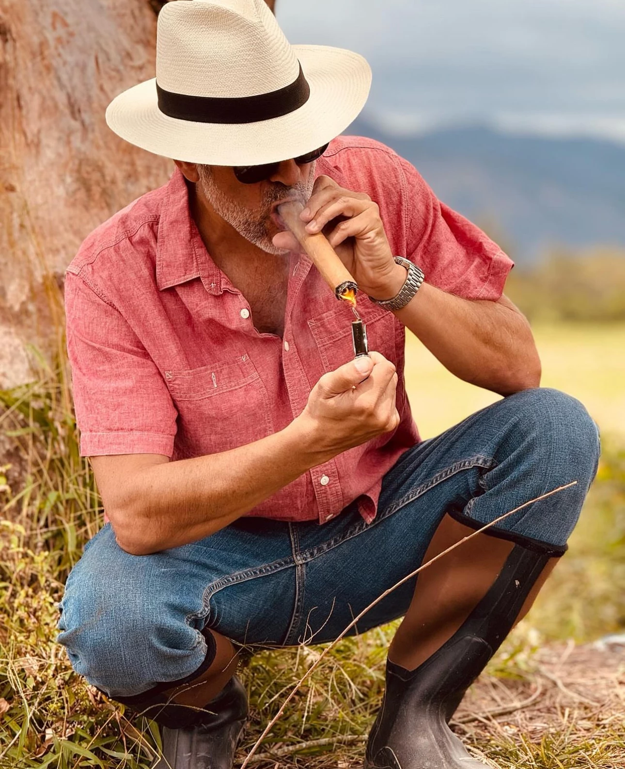 Iván Gancedo in a Panama hat, crouching by a tree and lighting a cigar with a torch lighter, in Jalapa