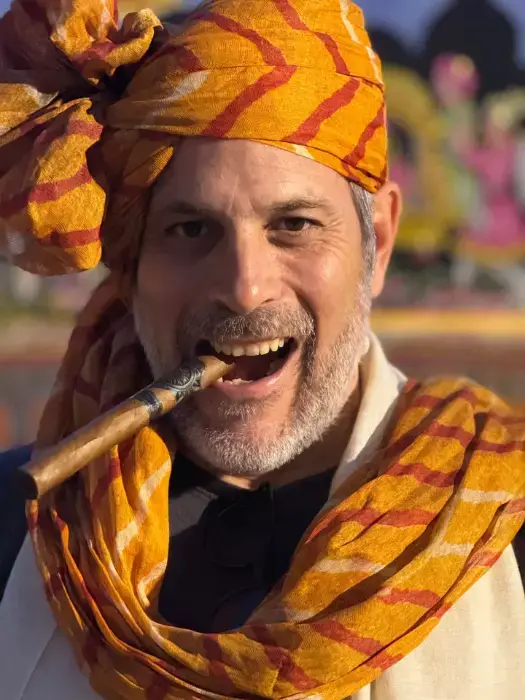 Portrait of Iván Gancedo in an orange patterned turban and scarf, cigar in his mouth and smiling, a colorful festival scene blurred behind him, in India