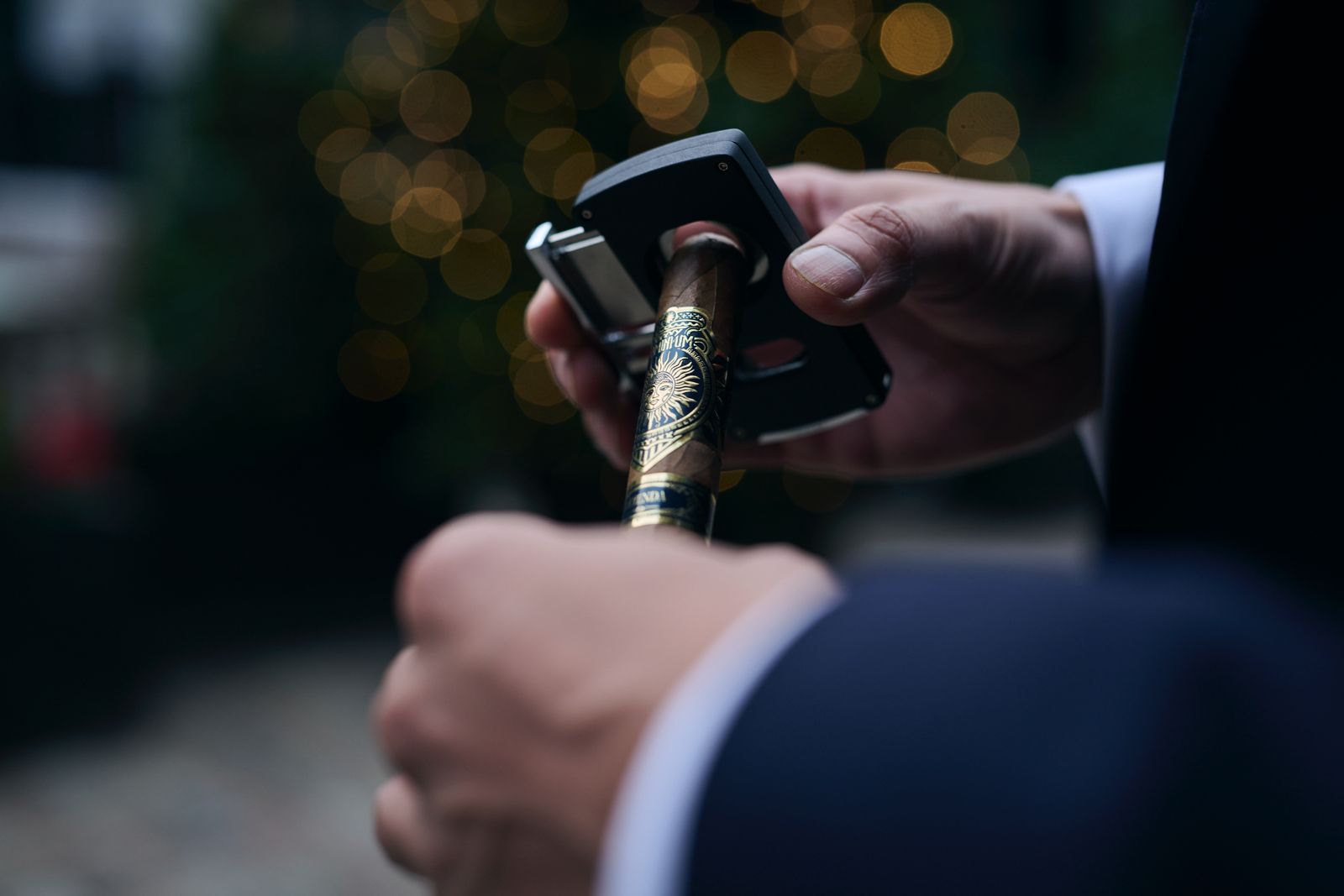 Close-up of hands cutting a Dominium cigar, string lights blurred behind in the evening