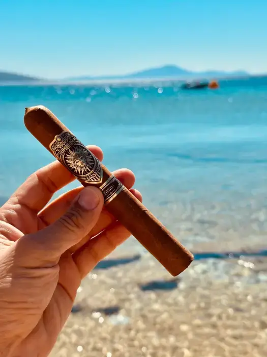 A hand holding a Dominium Leyenda cigar against a turquoise sea and beach